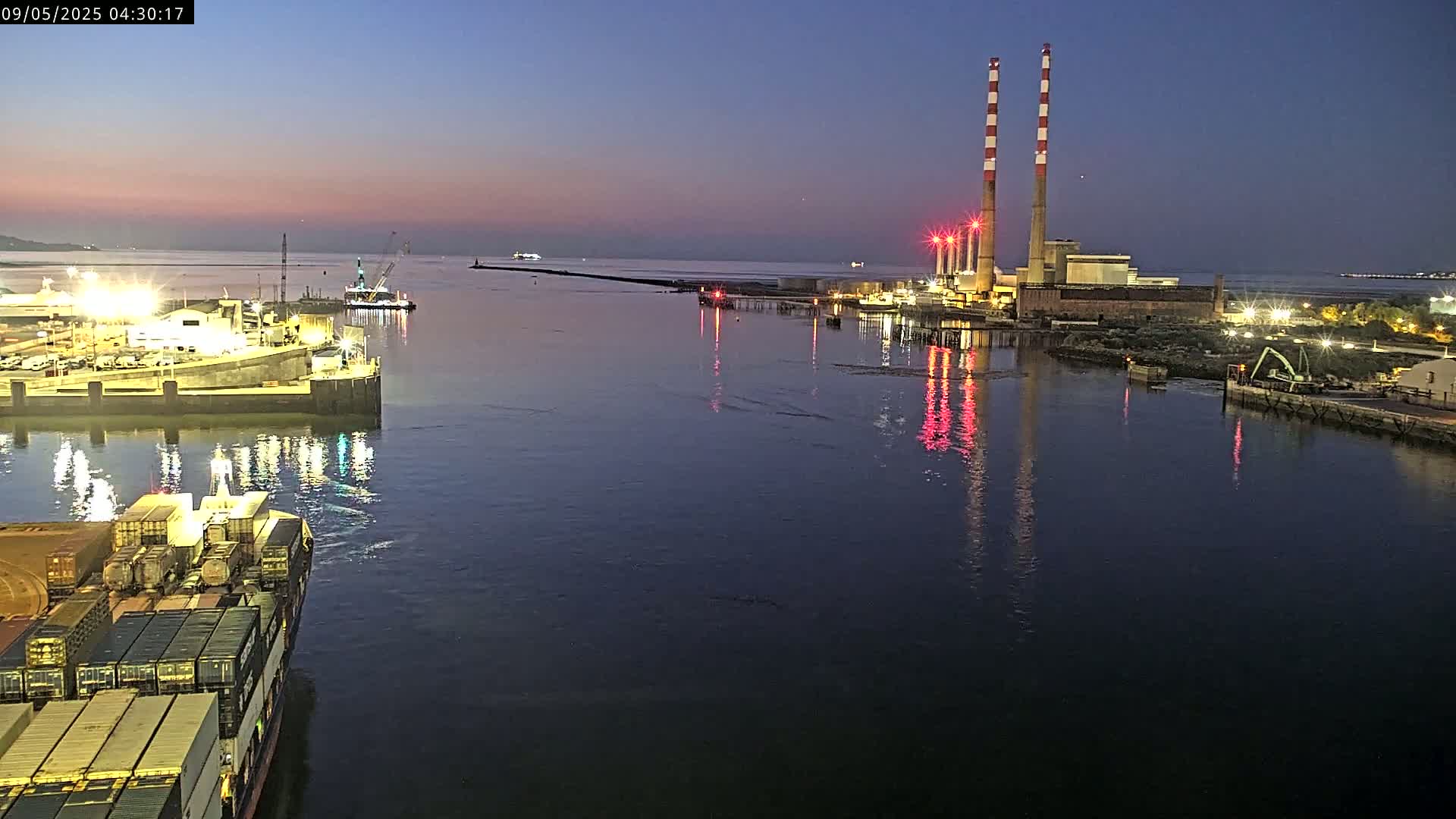 A container ship is in a harbor at twilight, with a power plant and other vessels visible in the distance.