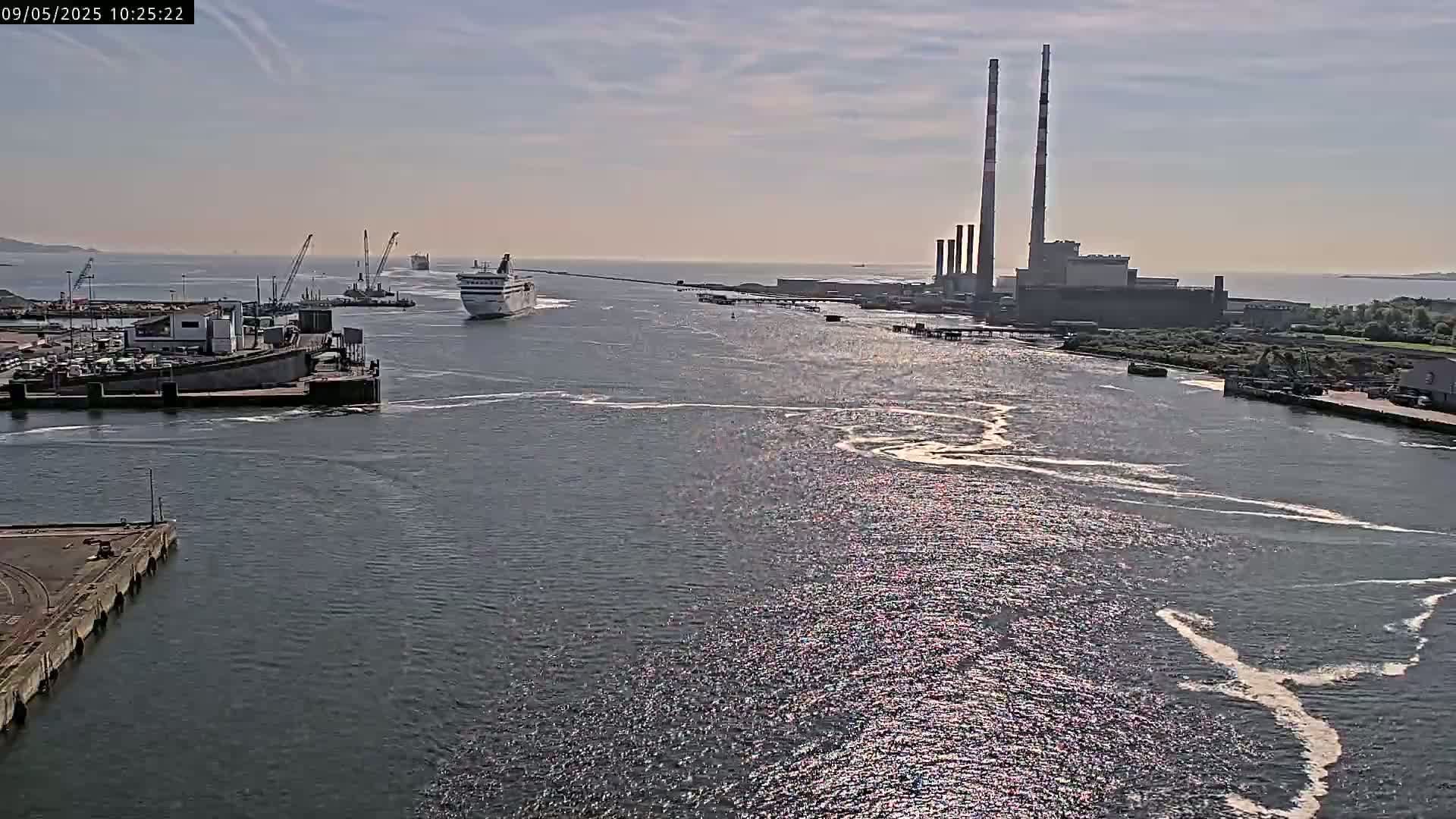 A ferry approaches a port under a mostly sunny sky, passing by a large industrial facility with tall smokestacks.