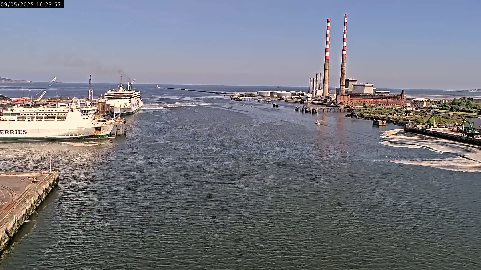 Two ferries are docked near a power plant with two tall smokestacks on a sunny day.