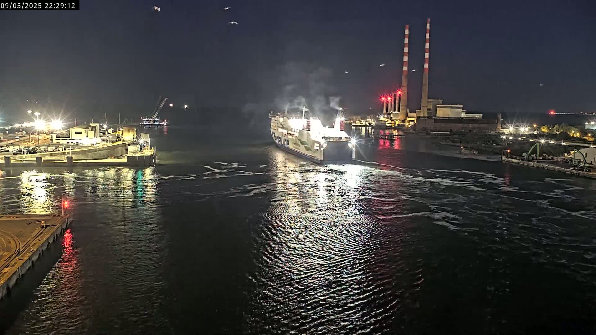 A large ferry approaches a dock at night under a clear, dark sky, with lights illuminating the waterway and surrounding industrial structures.