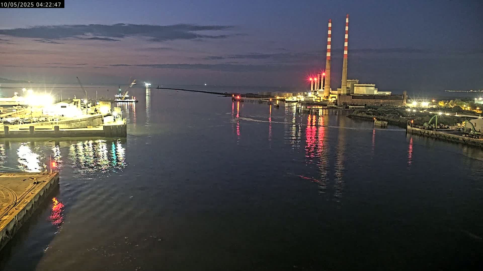 A nighttime view of a harbor, illuminated by lights from docked ships and a power plant with tall chimneys, under a dark sky.