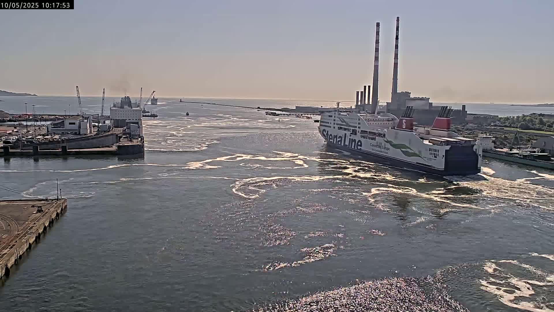 A large ferry departs a harbor on a sunny day, leaving a wake in its path, with other ships and industrial structures visible in the background.