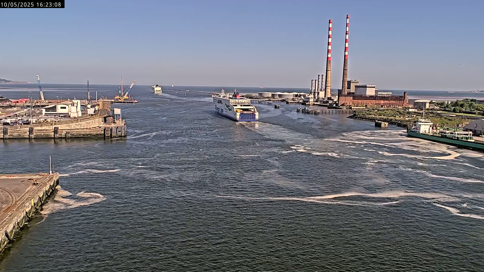 A ferry navigates a harbor on a sunny day, passing near industrial structures with tall smokestacks.