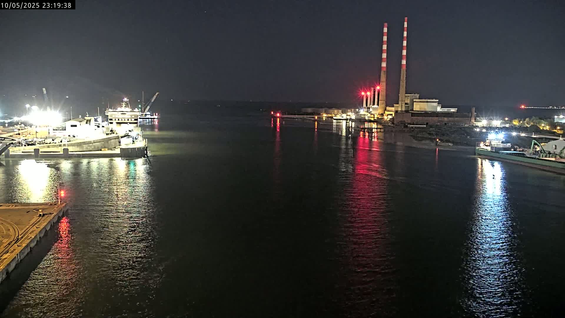 At night, a body of water reflects the lights of docked ships and a power plant with tall smokestacks.