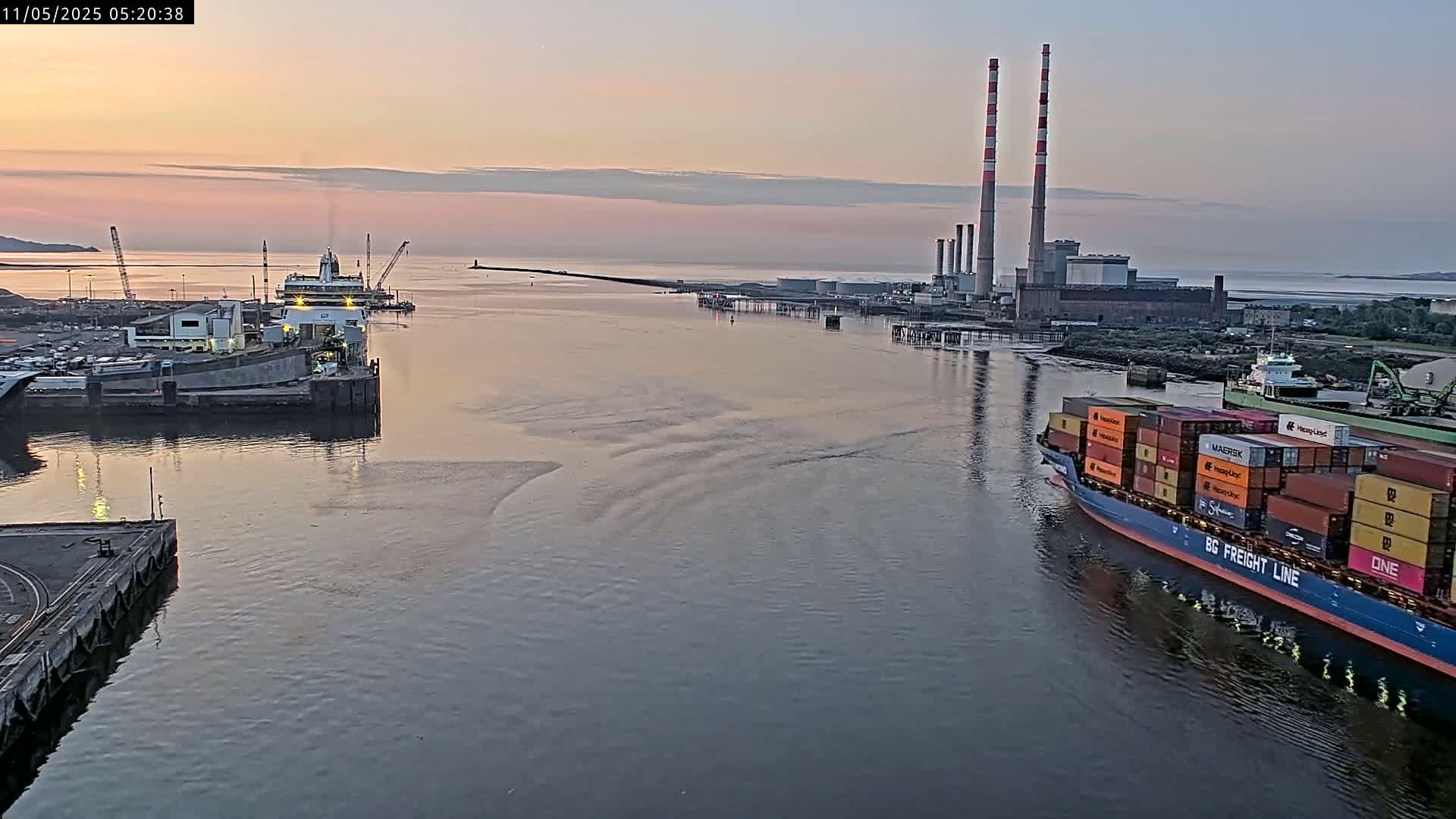 A container ship sails past a ferry docked at a harbor during a calm sunrise, with a power plant visible in the background.
