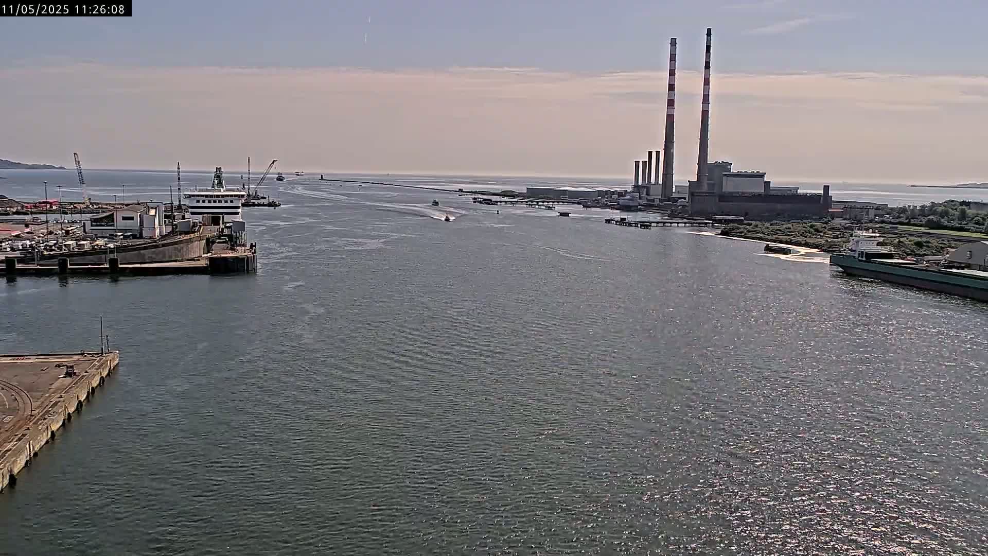 A wide shot of a harbor on a sunny day shows several ships and a power plant in the distance.