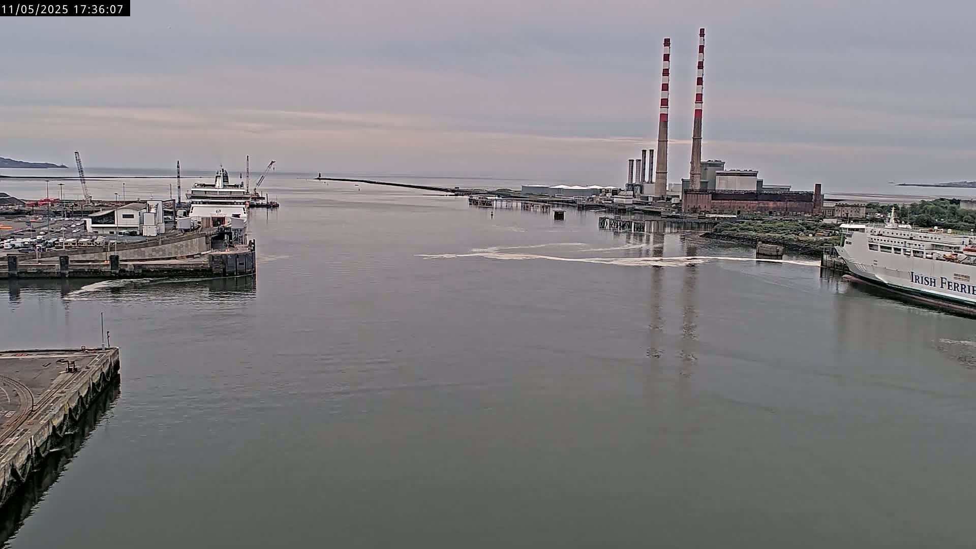A calm, overcast day reveals two ferries docked at a port near an industrial facility with tall smokestacks.