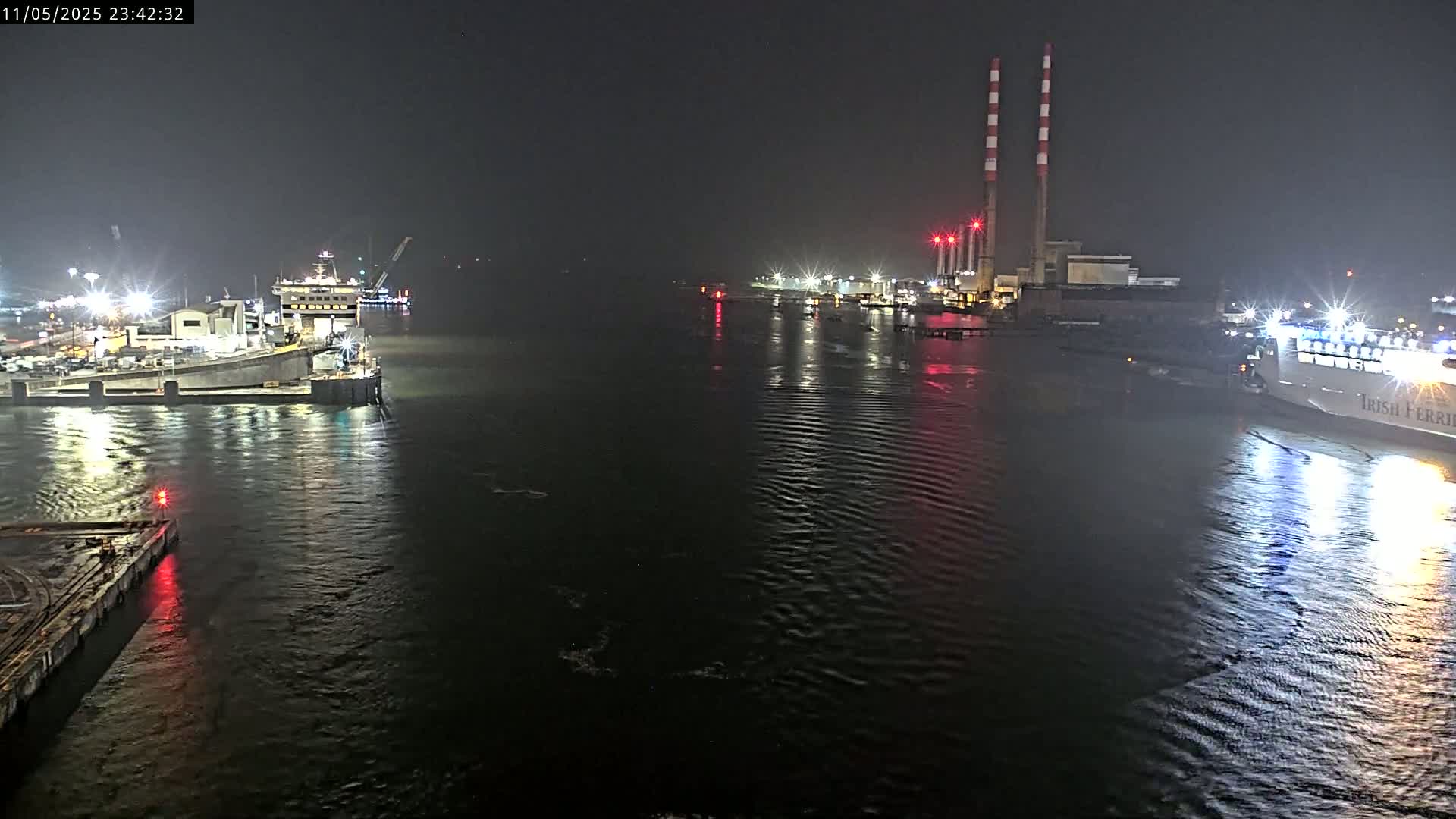 At night, a dark body of water reflects the lights of docked ships and a power plant on its shore.