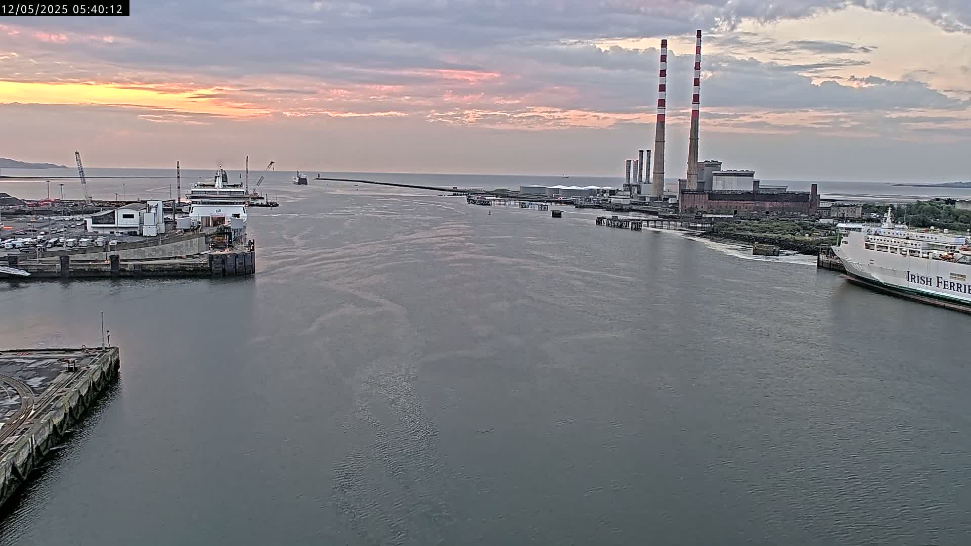 A calm harbor at sunrise shows two ferries docked near a power plant with tall smokestacks.