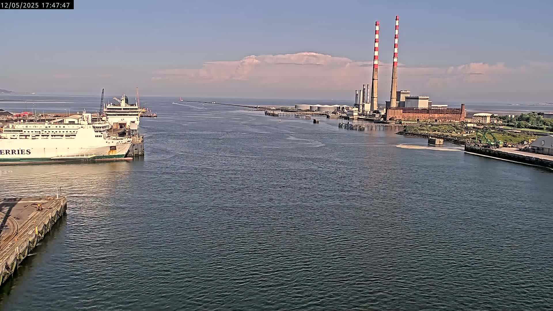 A ferry is docked at a port next to a power plant with two tall smokestacks under a mostly clear sky.