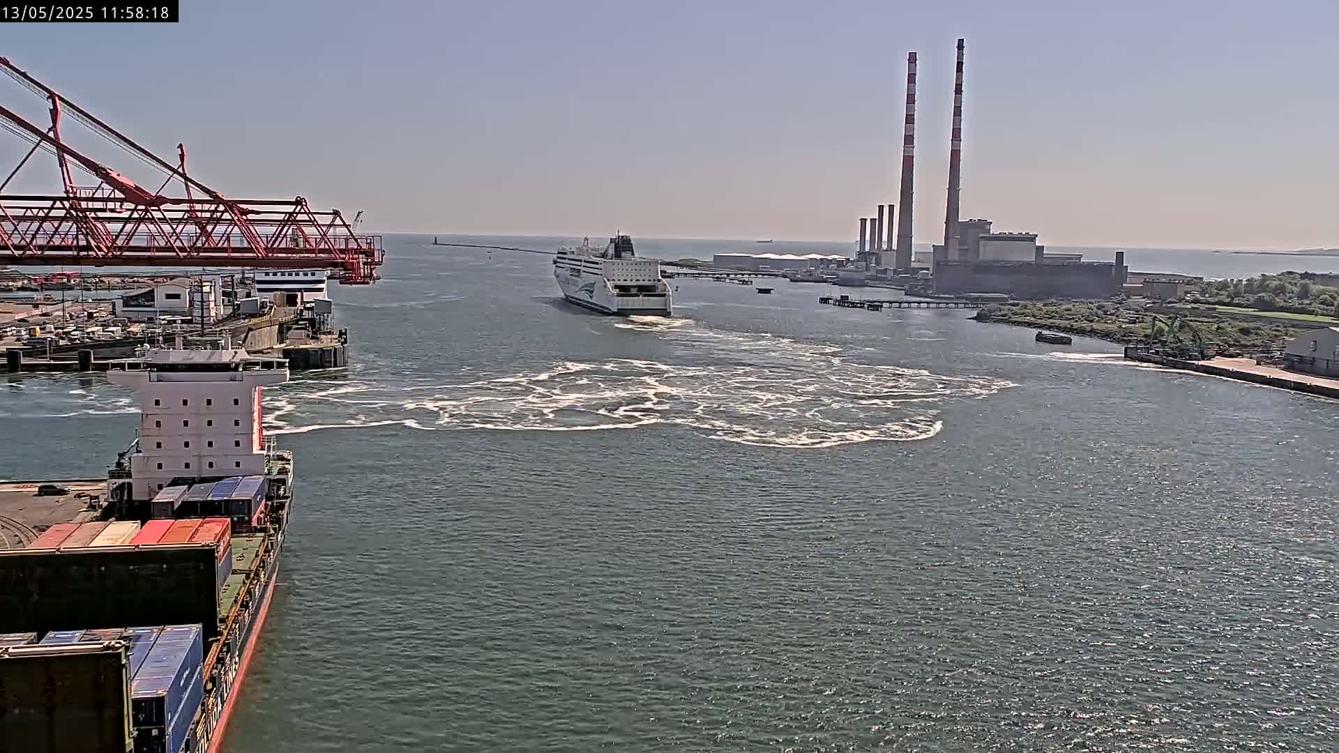 A ferry passes a container ship in a harbor on a sunny day, with a large industrial plant visible in the background.