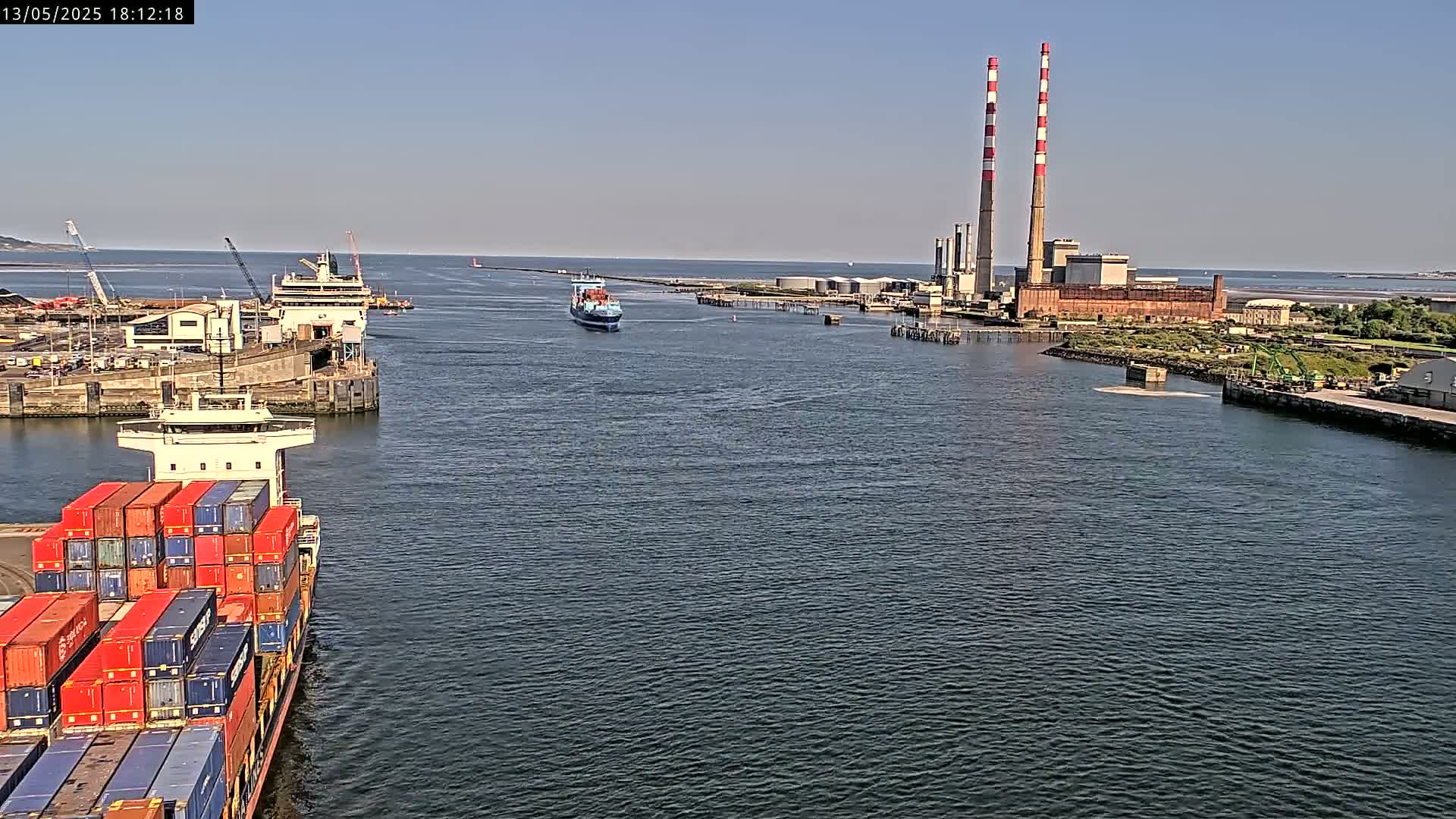 A container ship sails past a docked ferry, toward a power plant with two tall smokestacks on a sunny day.