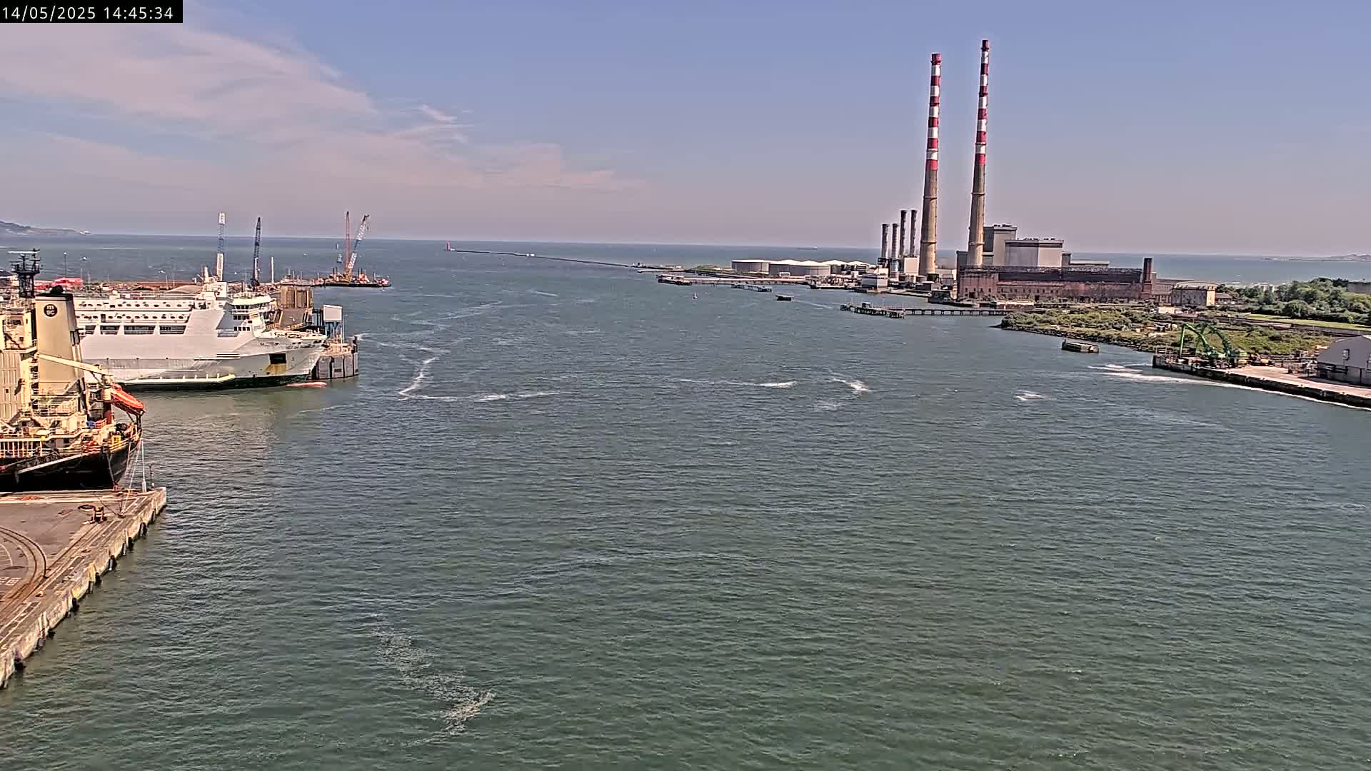 A harbor scene on a sunny day shows a ferry and cargo ship docked, with a power plant and tall smokestacks in the distance.