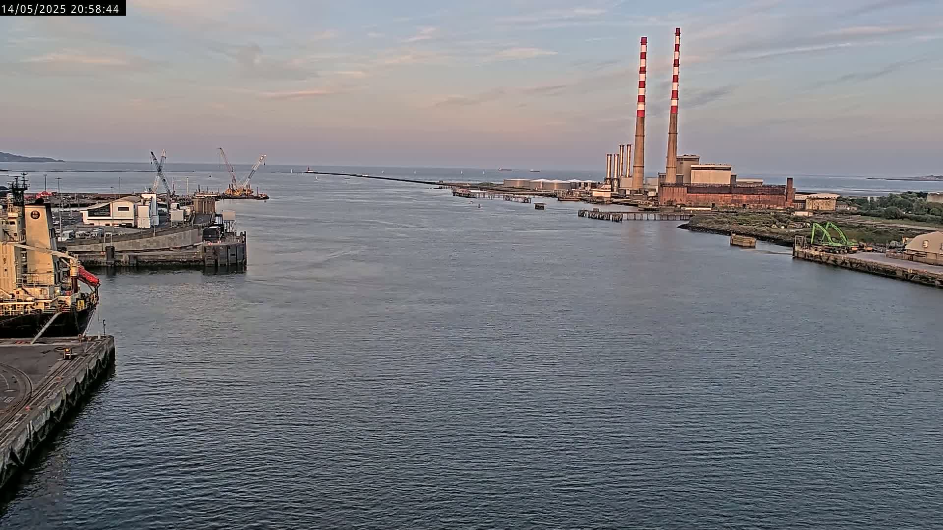 A calm body of water shows a cargo ship docked near an industrial area with tall smokestacks under a partly cloudy sky.