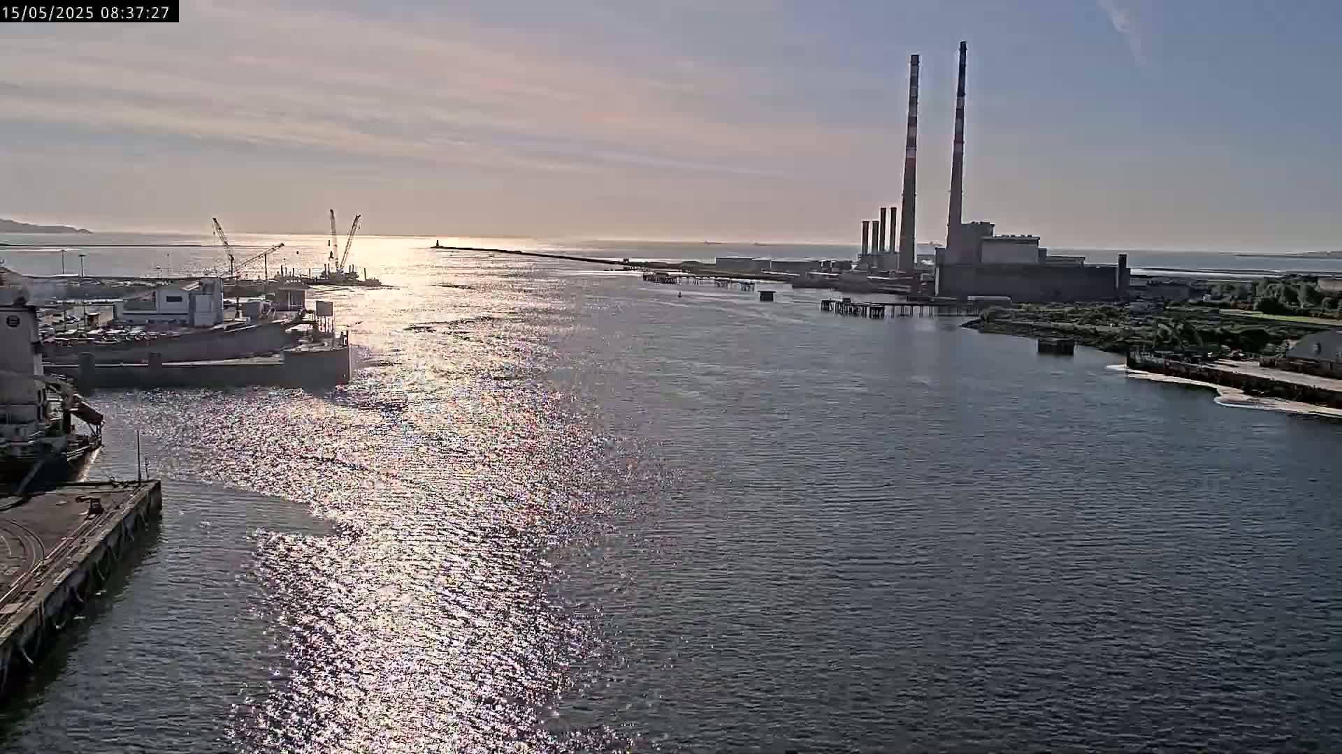 A wide, sunlit waterway shows a harbor with docked ships and cranes, a power plant with tall smokestacks in the distance, and calm water reflecting the light.