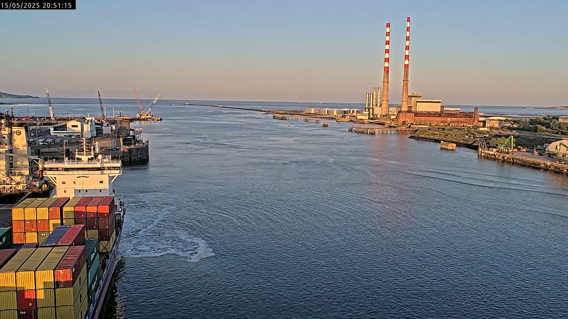 A container ship sails past a harbor at sunset, toward an industrial plant with tall smokestacks.