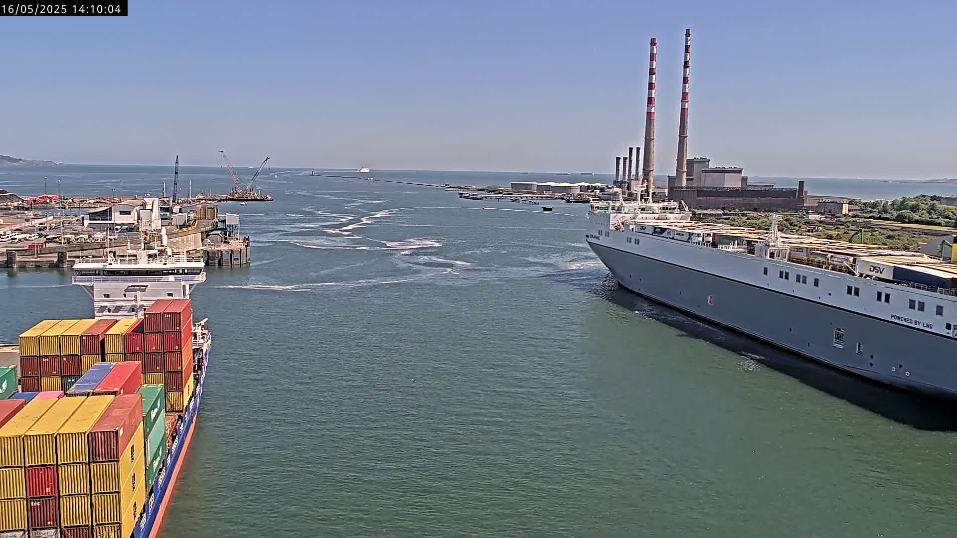 A container ship and a large cargo ship navigate a waterway on a sunny day, passing industrial buildings with tall smokestacks in the background.