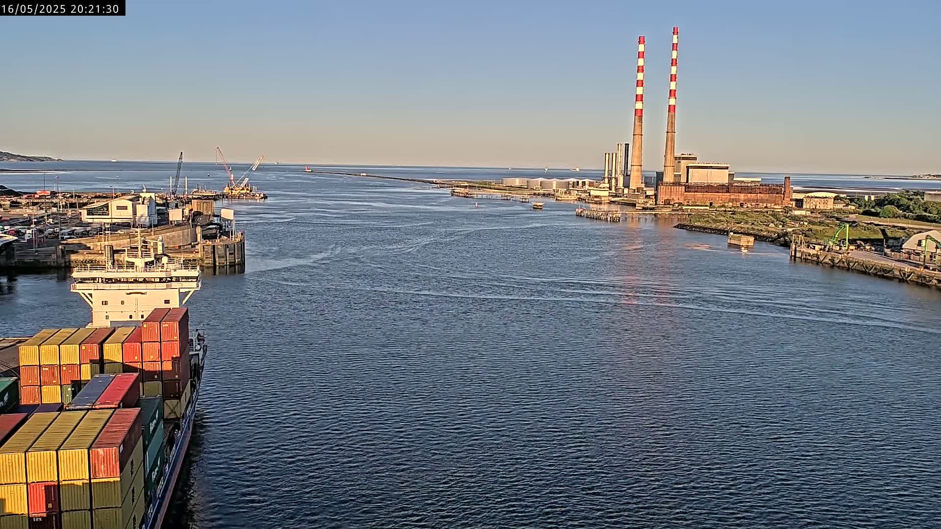 A container ship sails past an industrial waterfront under a clear, bright sky.
