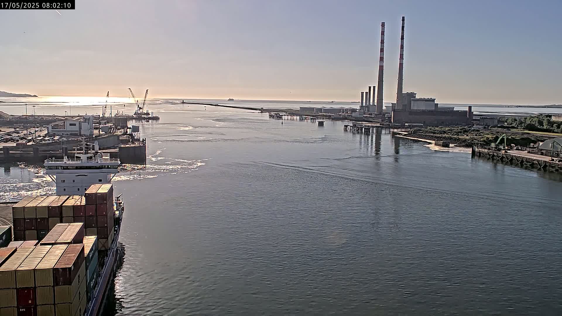 A container ship sails past a port facility with large industrial buildings and tall smokestacks under a clear, sunny sky.