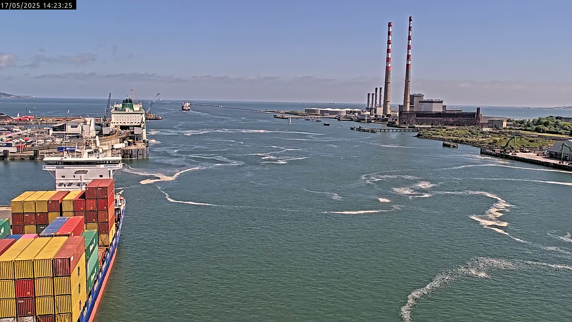 A container ship and a ferry are docked in a harbor on a sunny day, with two tall smokestacks and industrial buildings visible in the distance.
