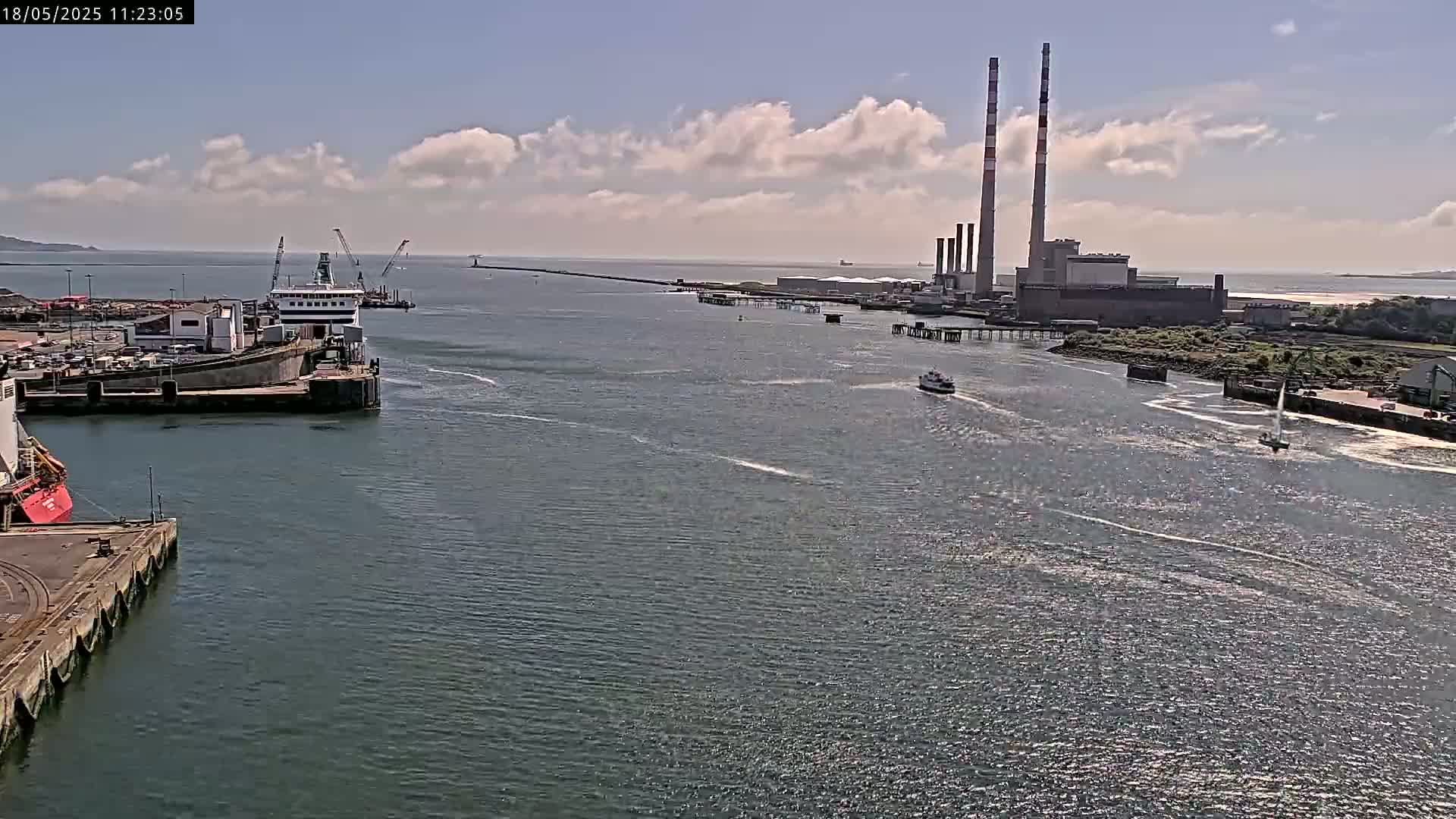 A ferry is docked at a port, with several smaller vessels navigating a waterway under partly cloudy skies, past a large industrial plant with tall smokestacks.