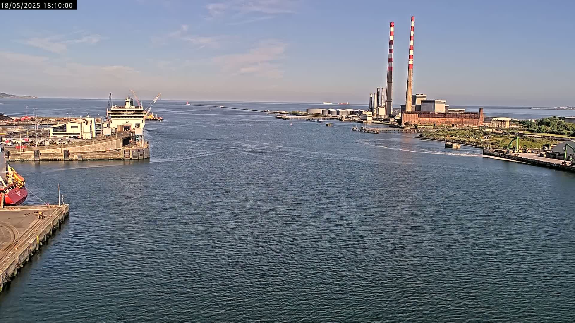 A wide shot of a harbor on a sunny day, showing a large ship docked near an industrial plant with two tall smokestacks.