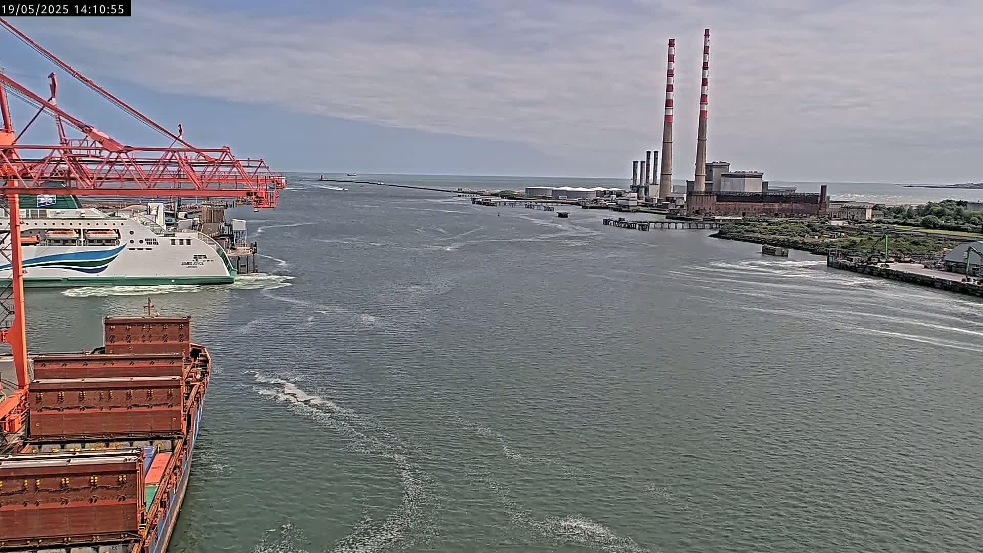 A large cargo ship and a ferry are in a harbor under a mostly cloudy sky, with a power plant and breakwater visible in the distance.