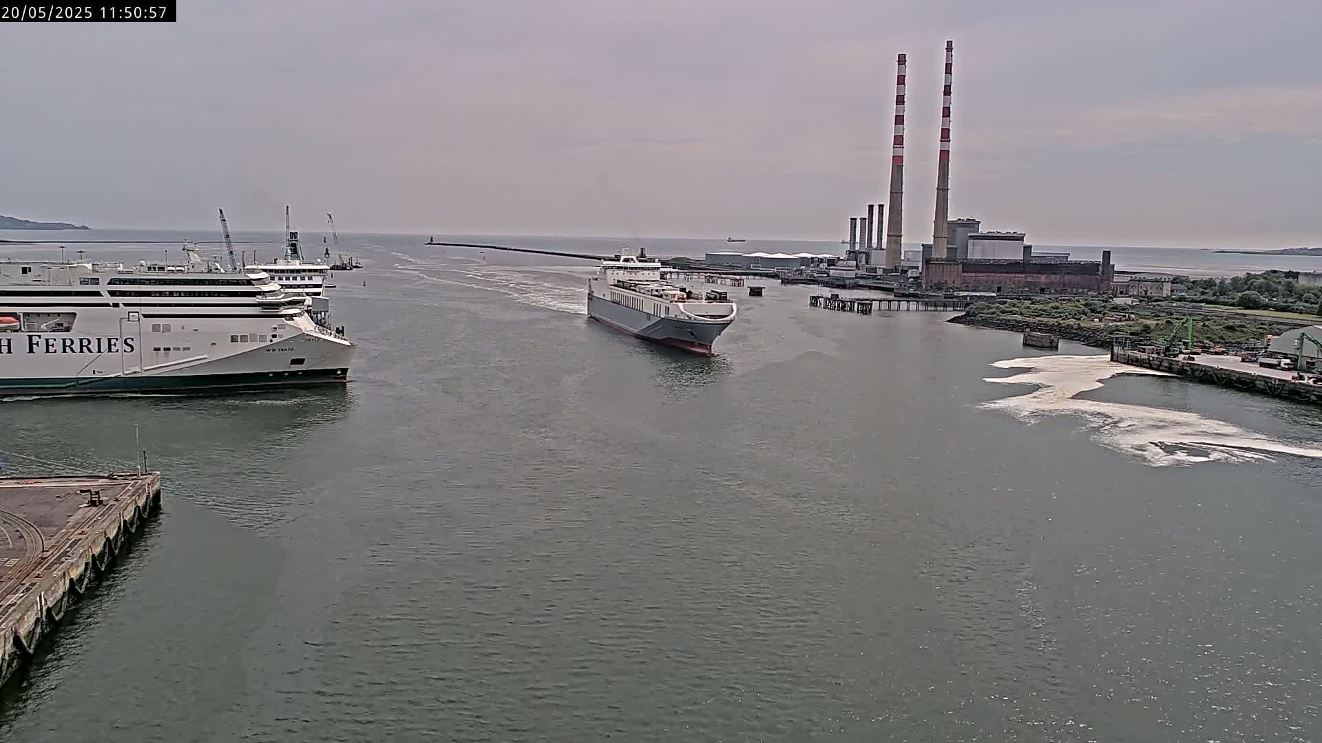 A large cargo ship and a ferry are seen navigating a waterway on an overcast day, with a power plant visible in the background.