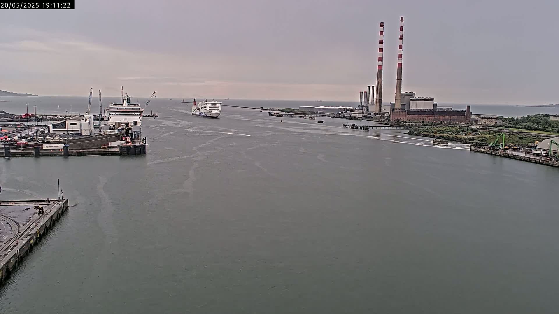 Two ferries are navigating a harbor toward a large industrial plant under an overcast sky.