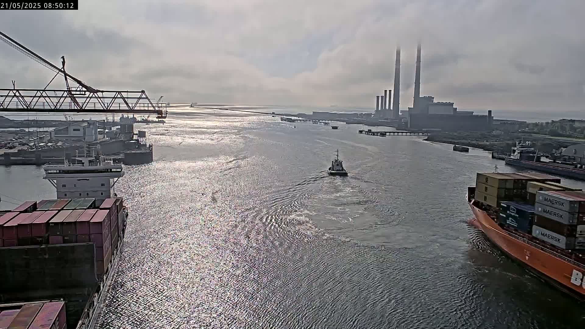 A wide shot of a waterway shows several ships, including container ships and a tugboat, with a power plant visible in the background under an overcast sky.