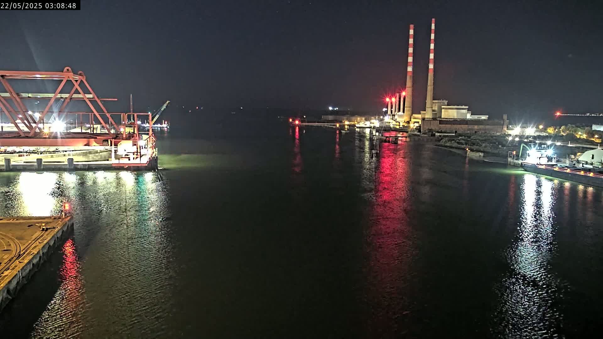 A nighttime view of a calm body of water shows a lit industrial area with tall smokestacks in the background, and a large piece of industrial equipment and a barge in the foreground.