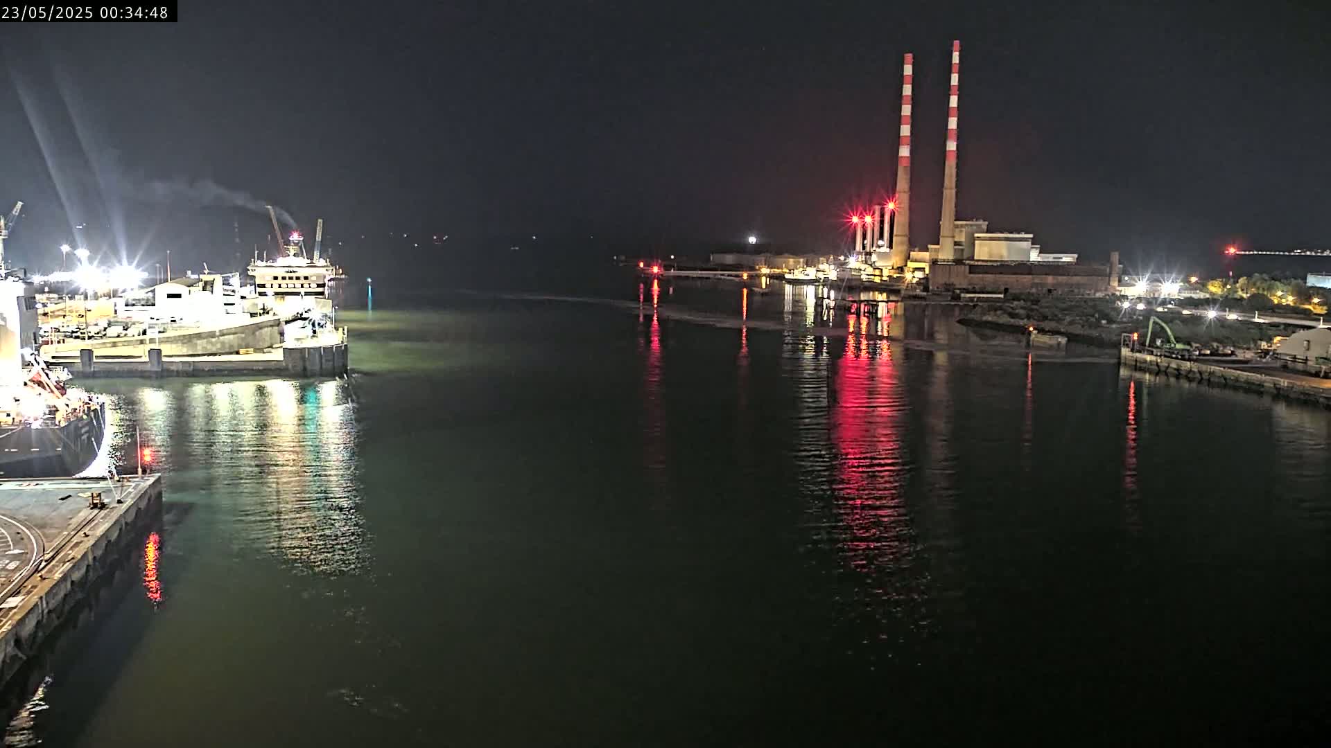A nighttime view of a harbor with docked ships and a power plant with tall smokestacks reflecting in calm water.