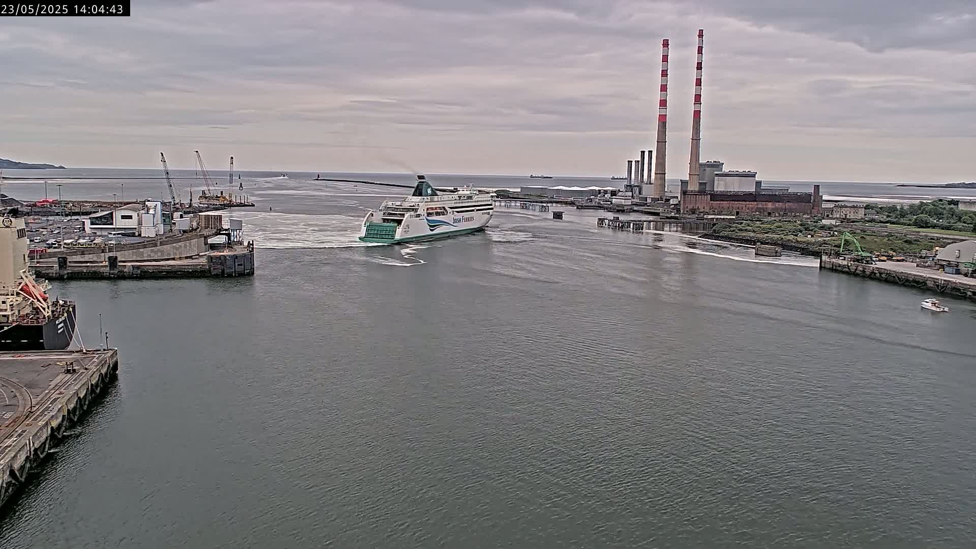 A ferry departs a harbor under an overcast sky, passing industrial buildings with tall smokestacks.