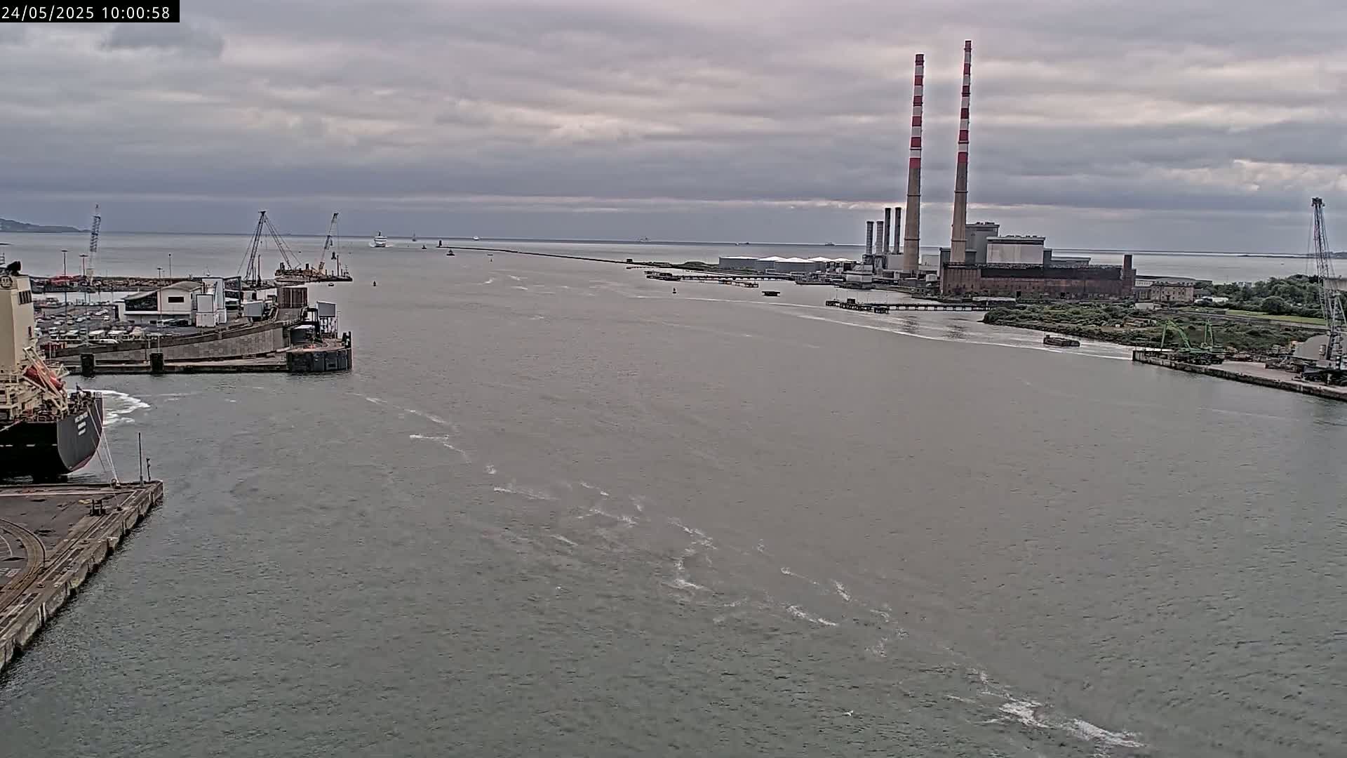 A wide, gray waterway shows a large industrial plant with tall smokestacks in the distance under an overcast sky, with various ships and docked vessels closer to the viewer.