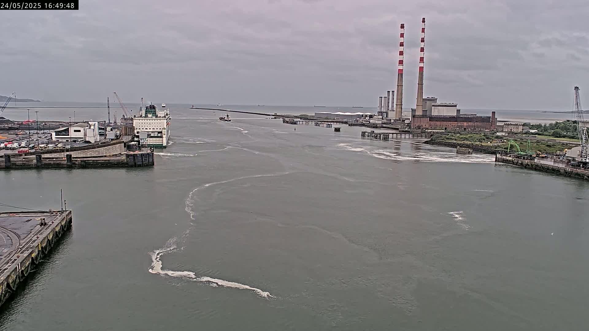 A ferry is docked at a harbor under an overcast sky, with a power plant visible in the distance.