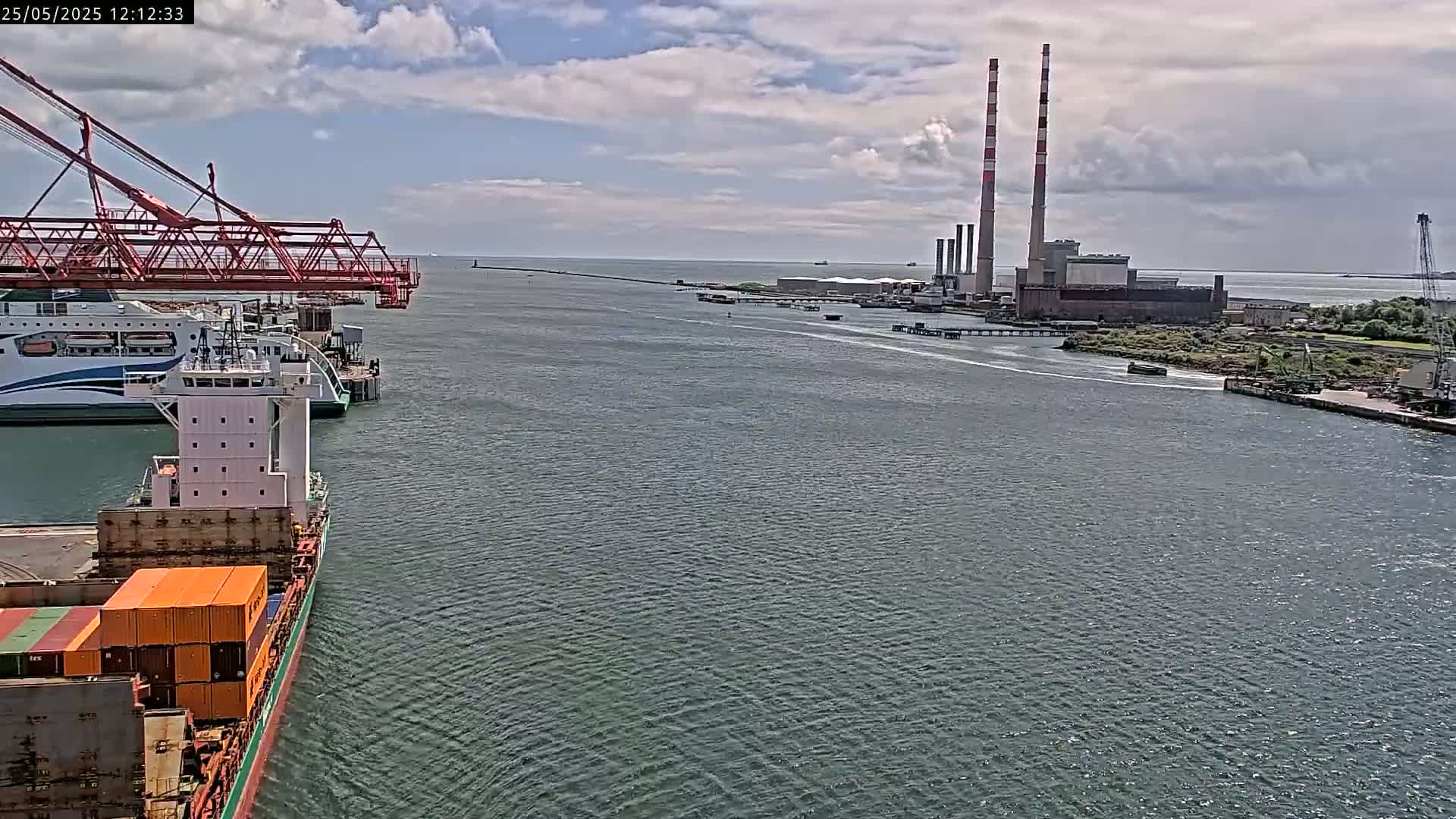 A container ship sails past a ferry and a large industrial plant with tall smokestacks on a partly cloudy day.