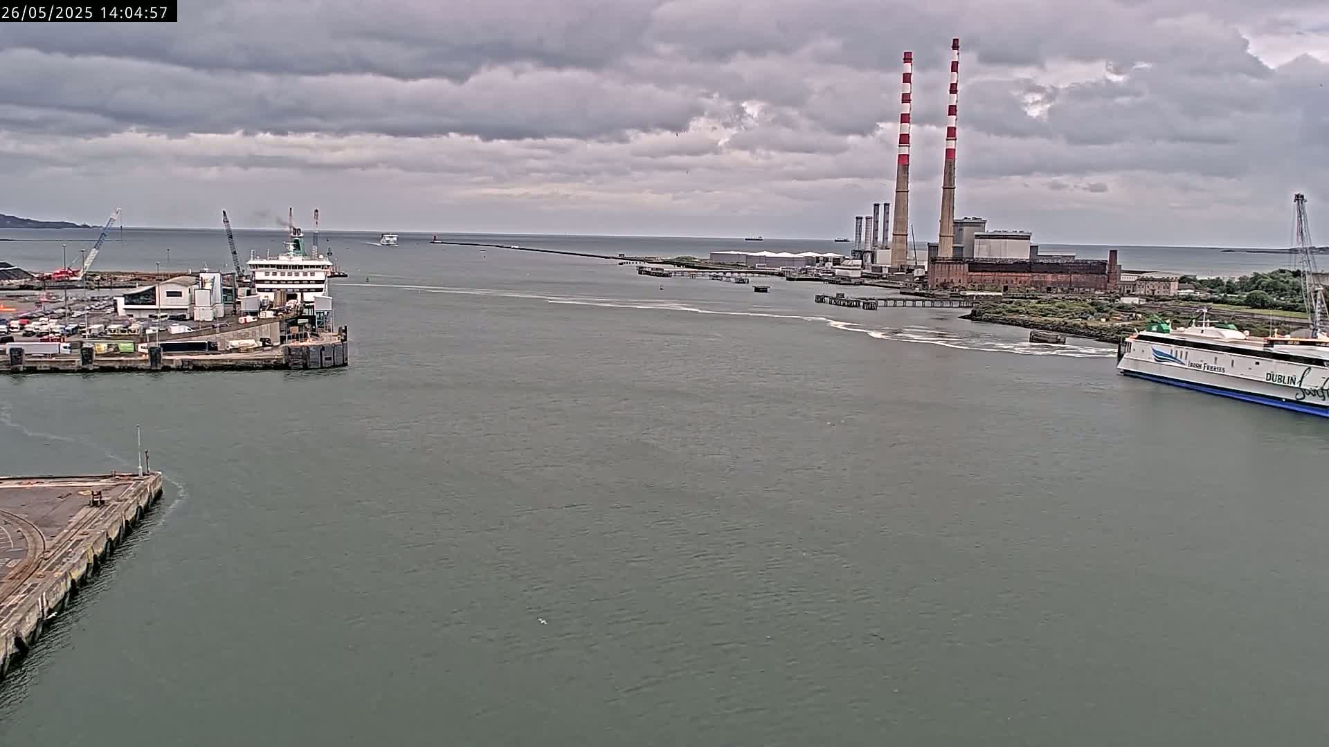 A ferry approaches a port under a cloudy sky, passing another ferry docked at a terminal near a power plant with tall smokestacks.