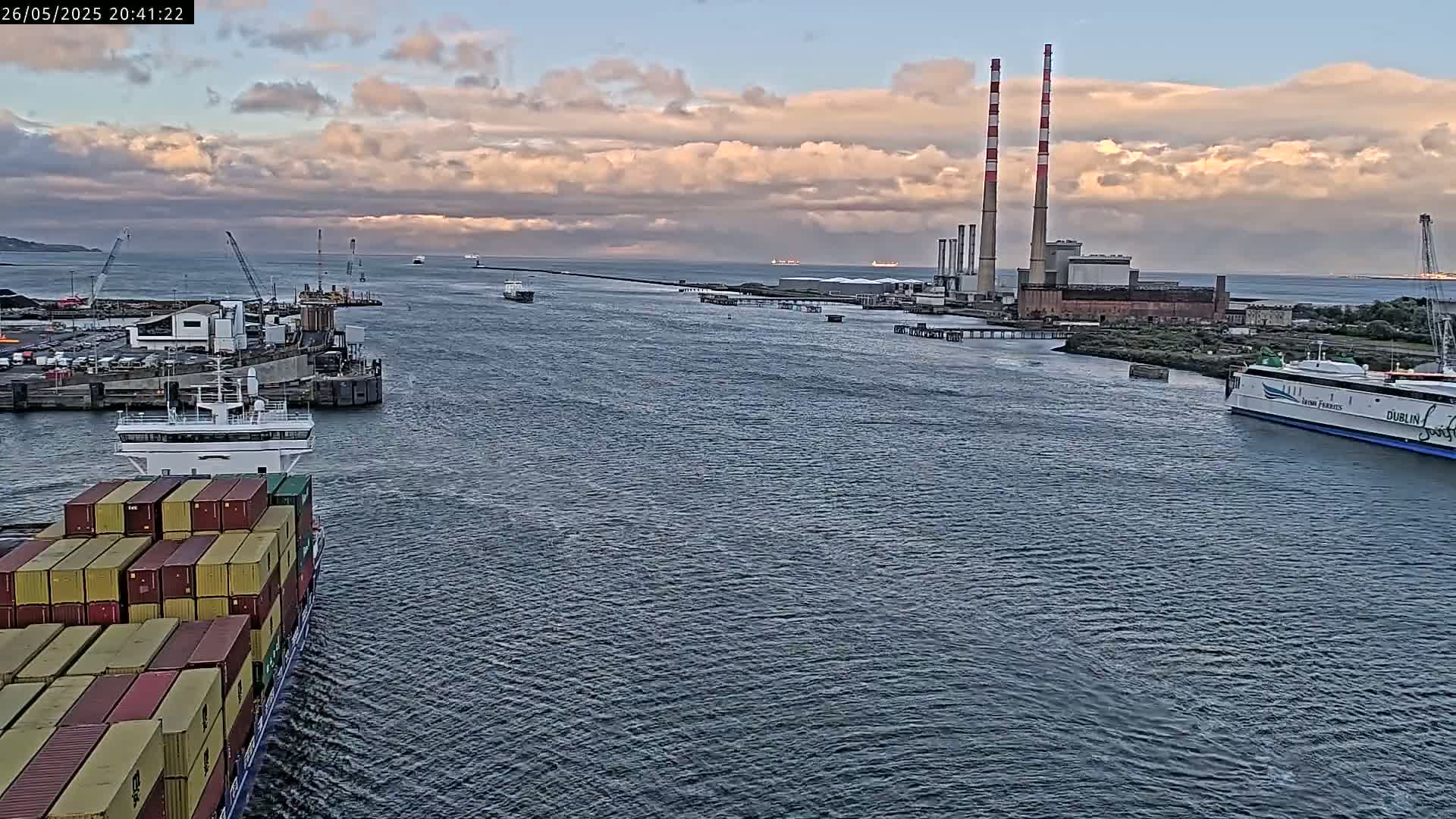 A container ship sails a waterway past a ferry and a power plant under a partly cloudy sky.
