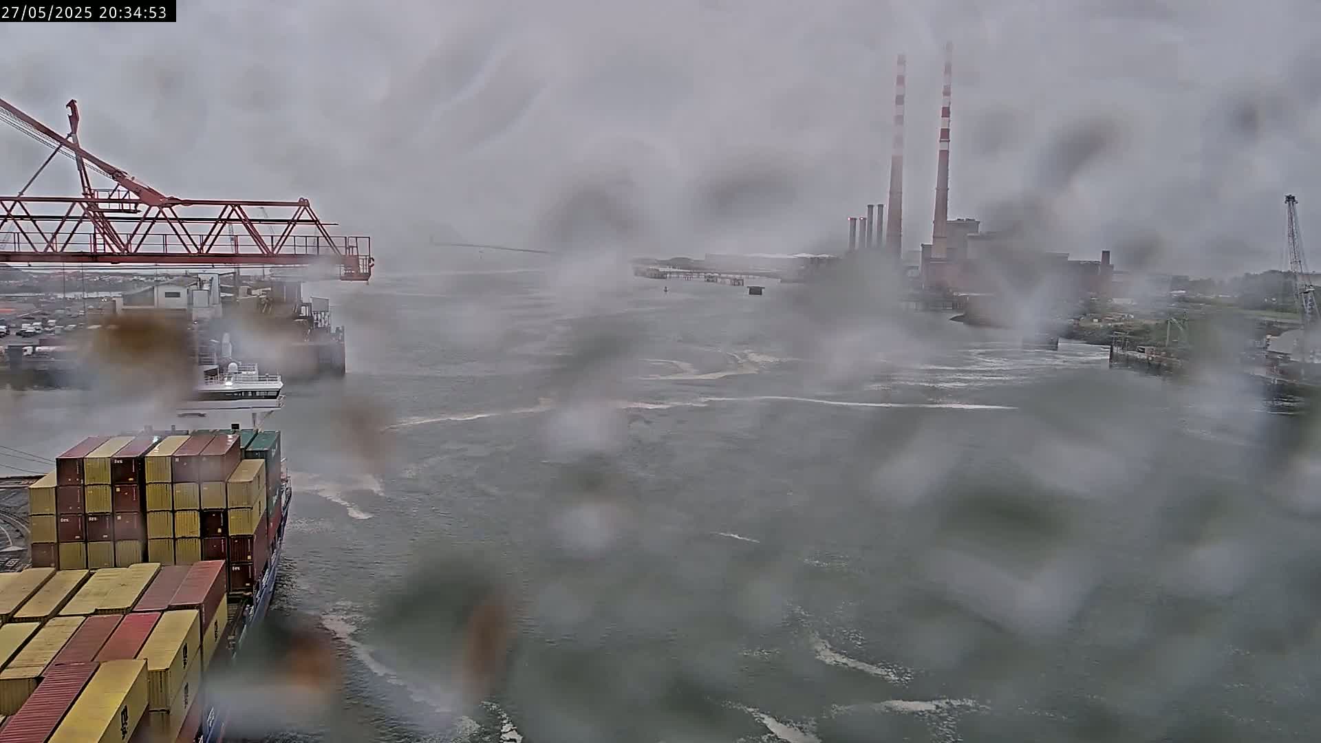 A rain-covered view of a container ship in a harbor, with a large industrial building and a crane visible in the distance.