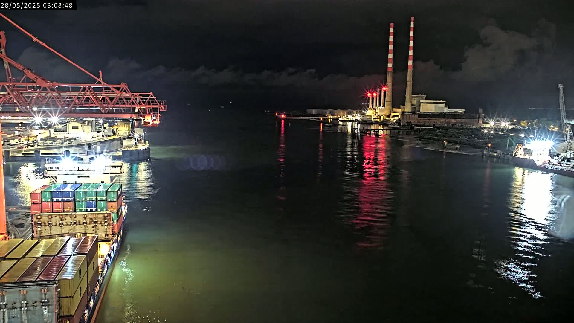 At night, a container ship is docked near a power plant with two tall smokestacks,  reflected in dark water under a partly cloudy sky.