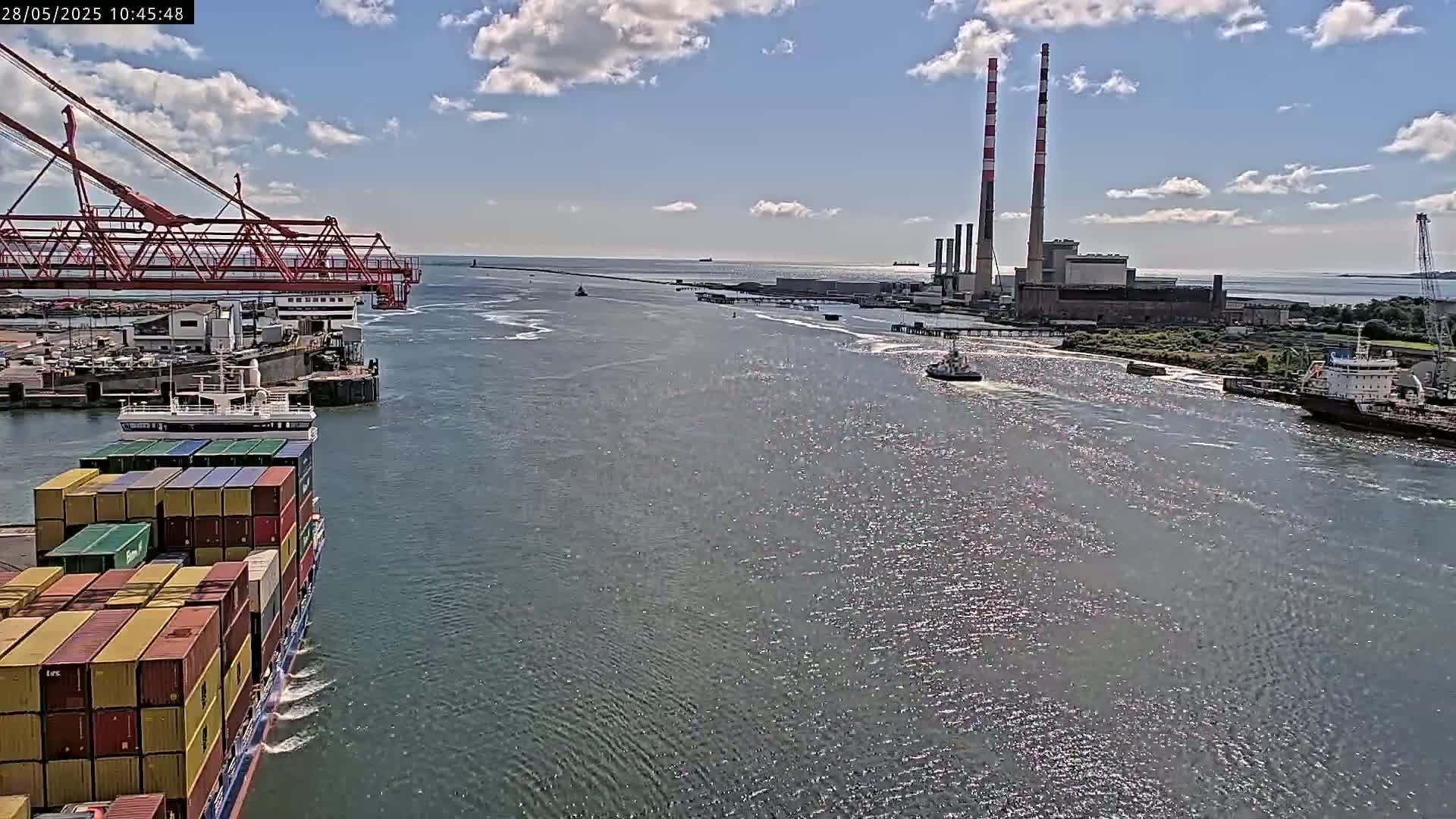 A container ship sails past a port under a partly cloudy sky, with a power plant and two tall smokestacks visible in the distance.
