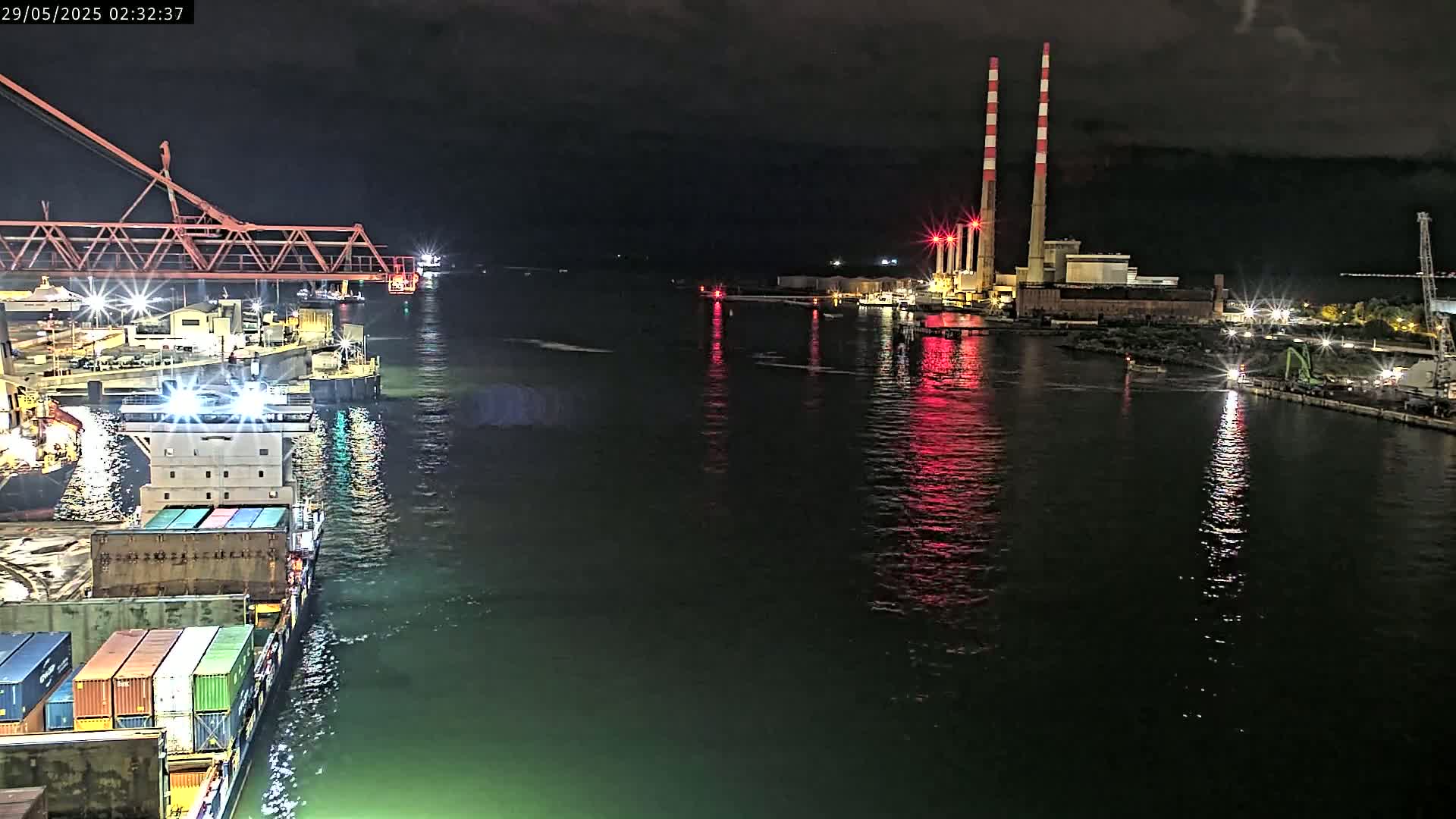 A container ship is docked at night near a power plant with tall red and white smokestacks, reflecting red light onto the dark water.