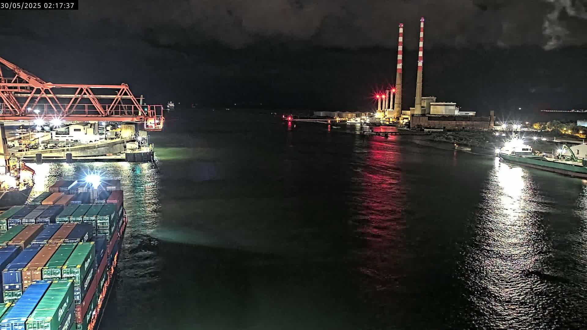 A container ship is docked at night next to a large industrial facility with tall smokestacks, illuminated by lights reflecting on dark water.