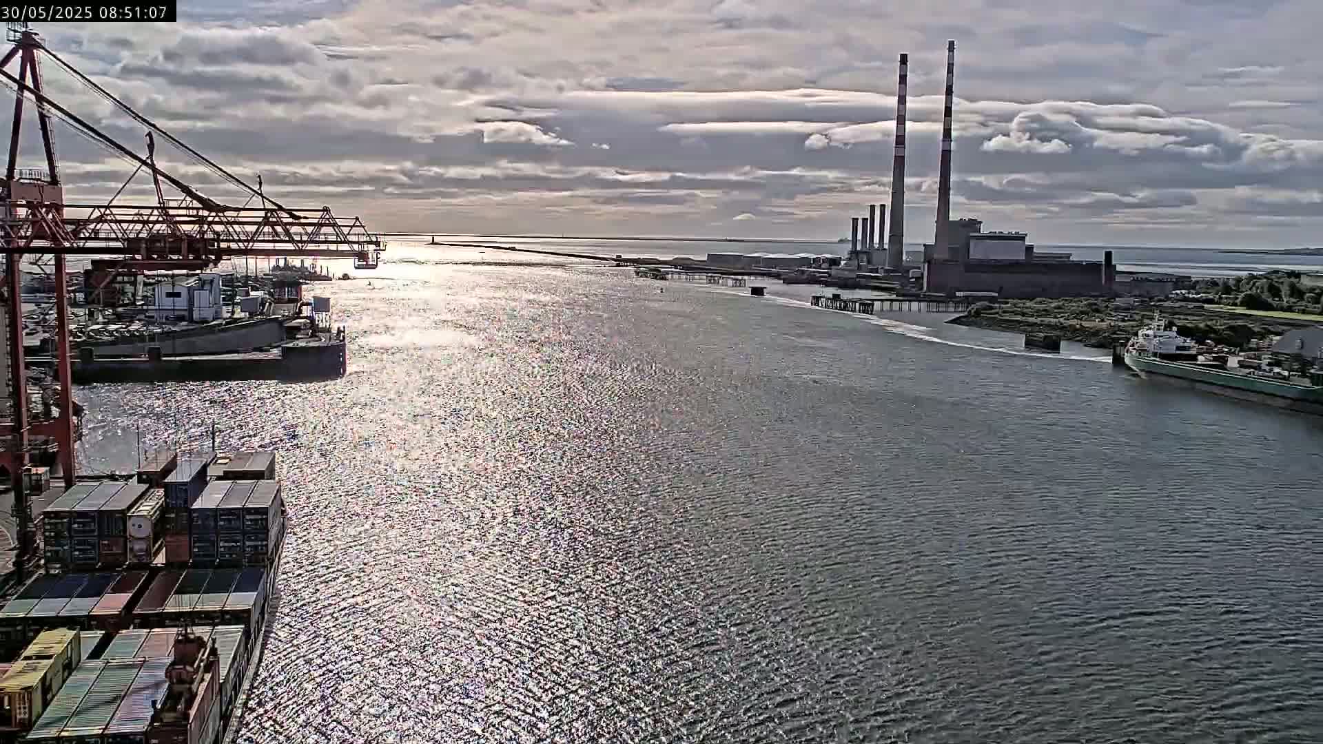 A partly cloudy day shows a wide waterway with container ships and a large industrial plant in the background.