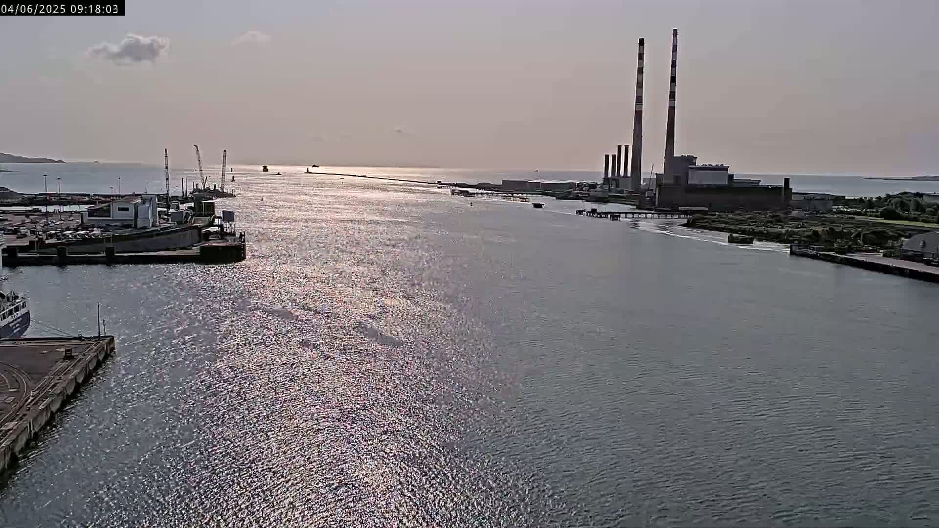 A calm, sunlit harbor scene shows a power plant with tall smokestacks in the background, several docked ships in the foreground, and a long jetty extending into the water.