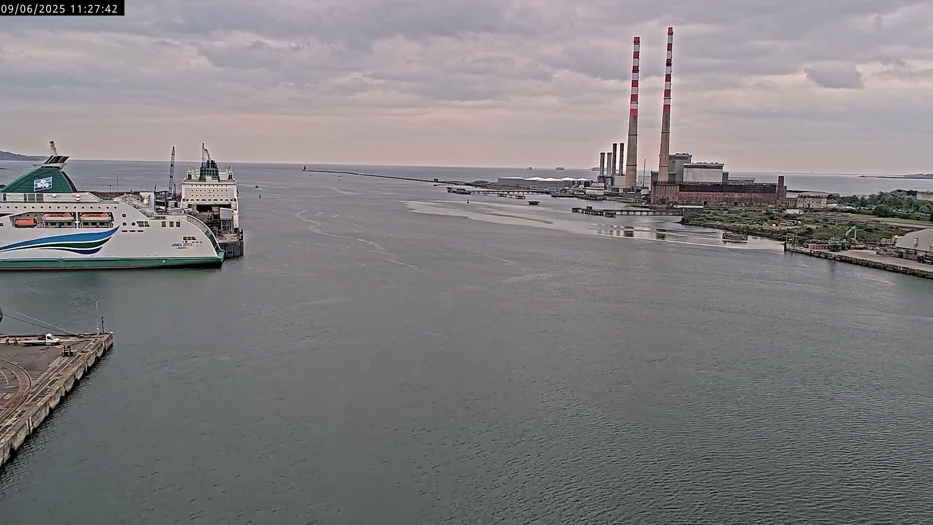 A ferry is docked at a port near an industrial facility with tall smokestacks, under a cloudy sky.