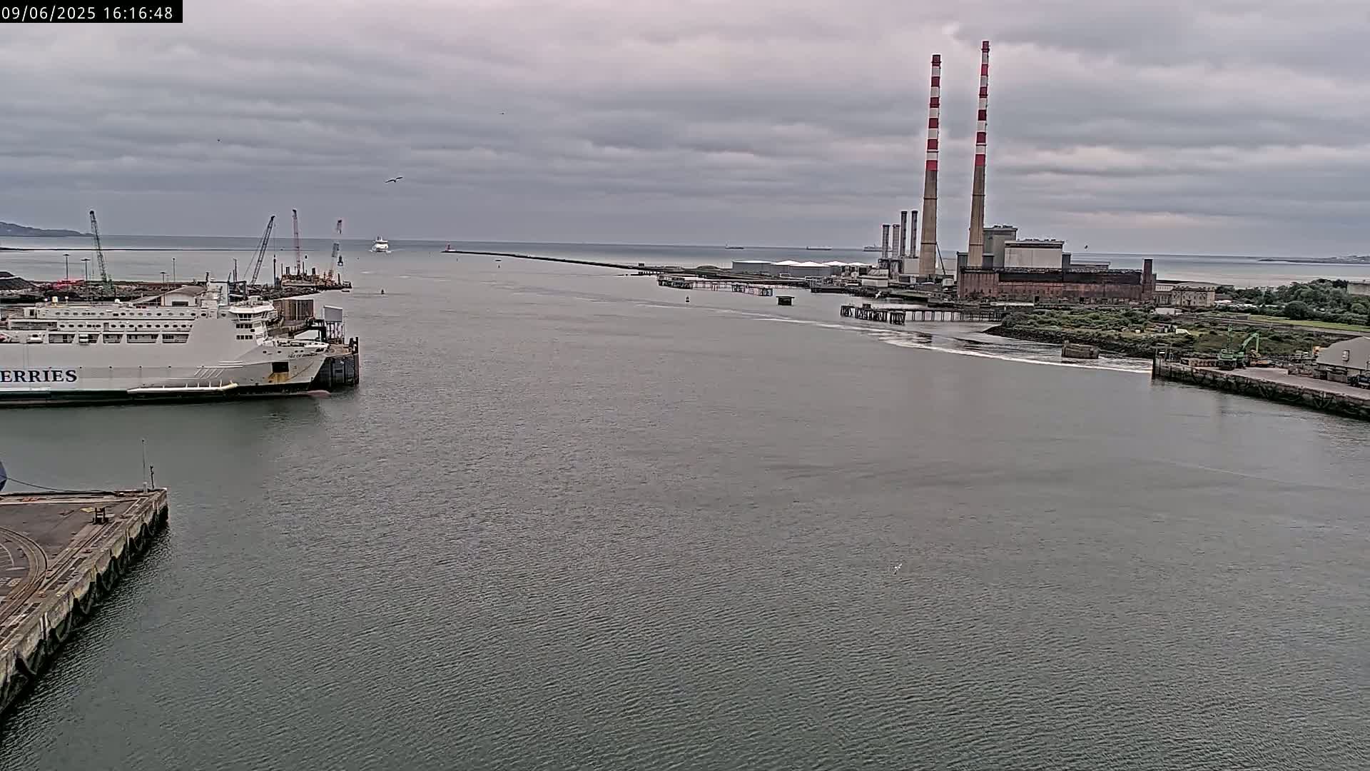 A ferry is docked at a harbor under a cloudy sky, with a power plant and breakwater visible in the distance.