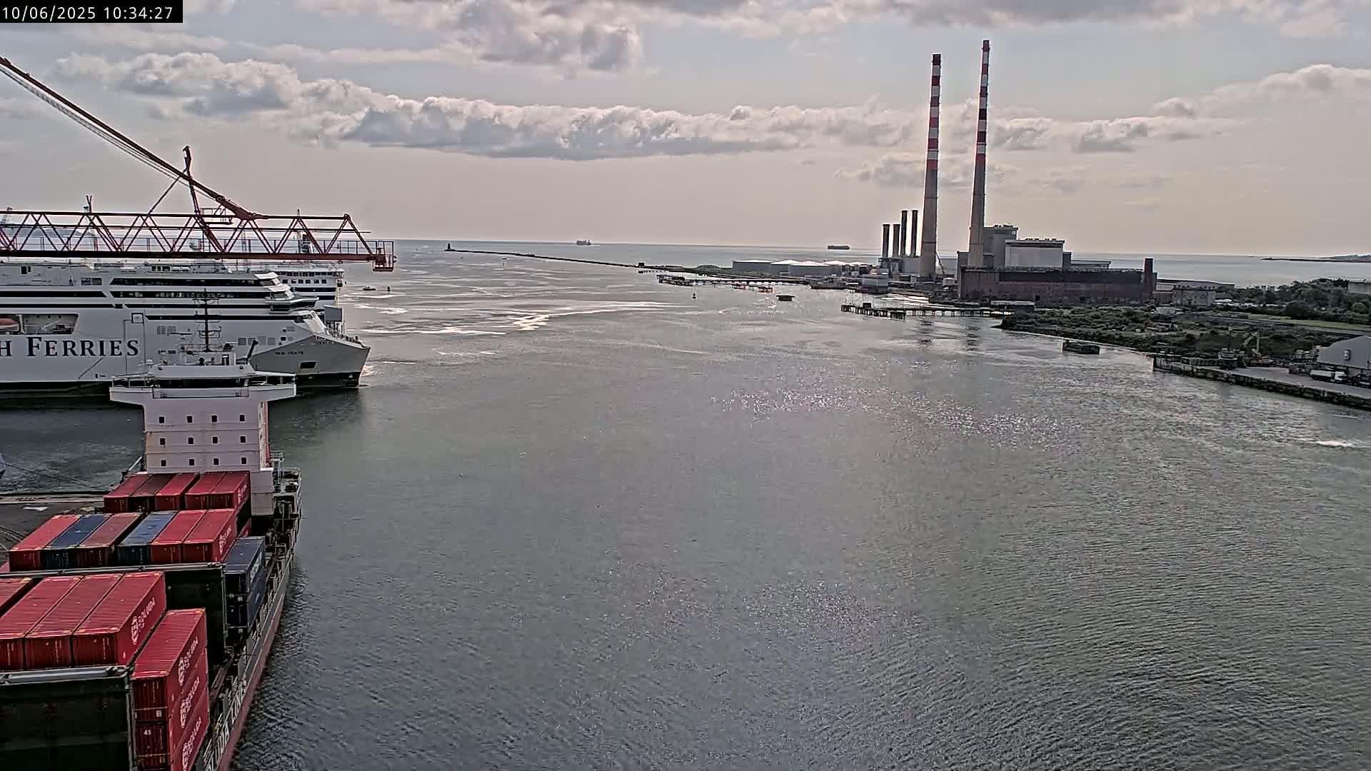 A container ship and a ferry are docked near an industrial plant with tall smokestacks under a partly cloudy sky.