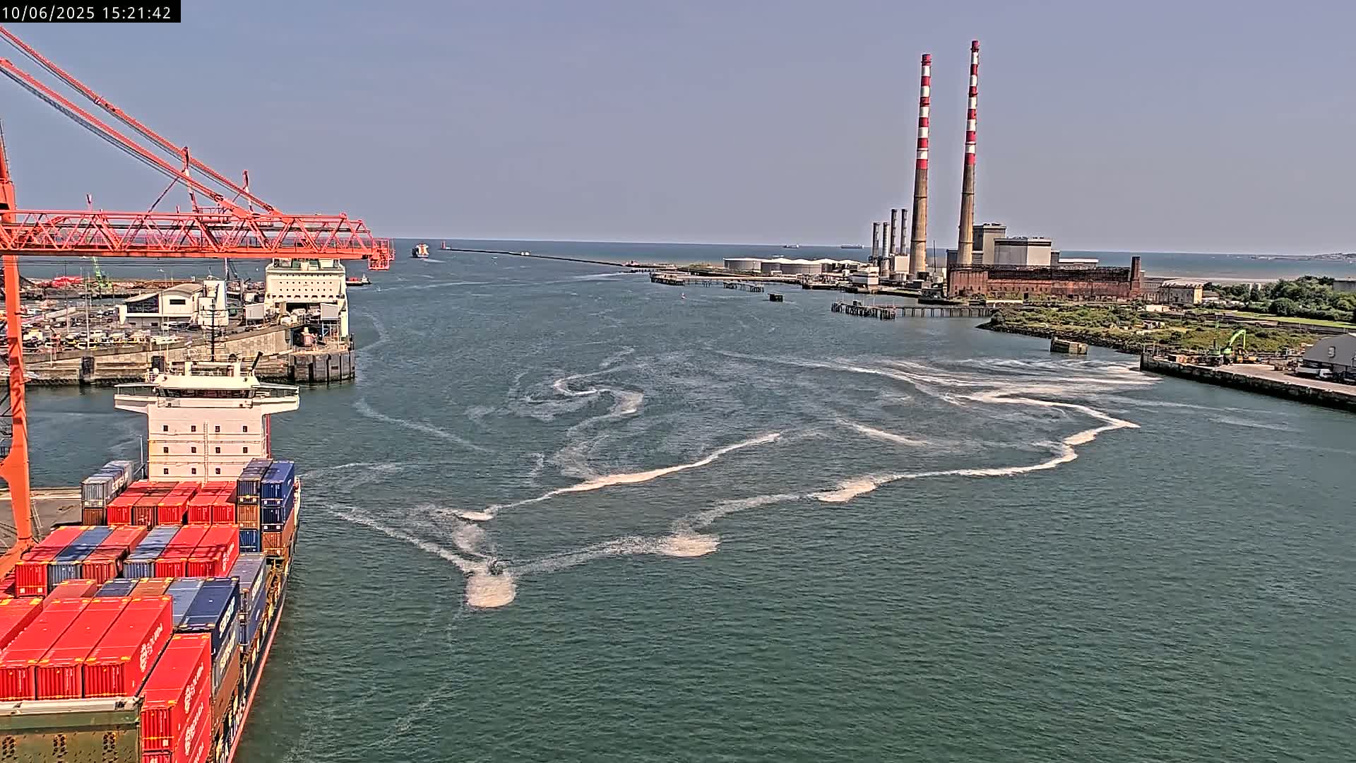 A container ship sails past a port on a sunny day, leaving a foamy wake in the water, with tall industrial chimneys visible in the distance.