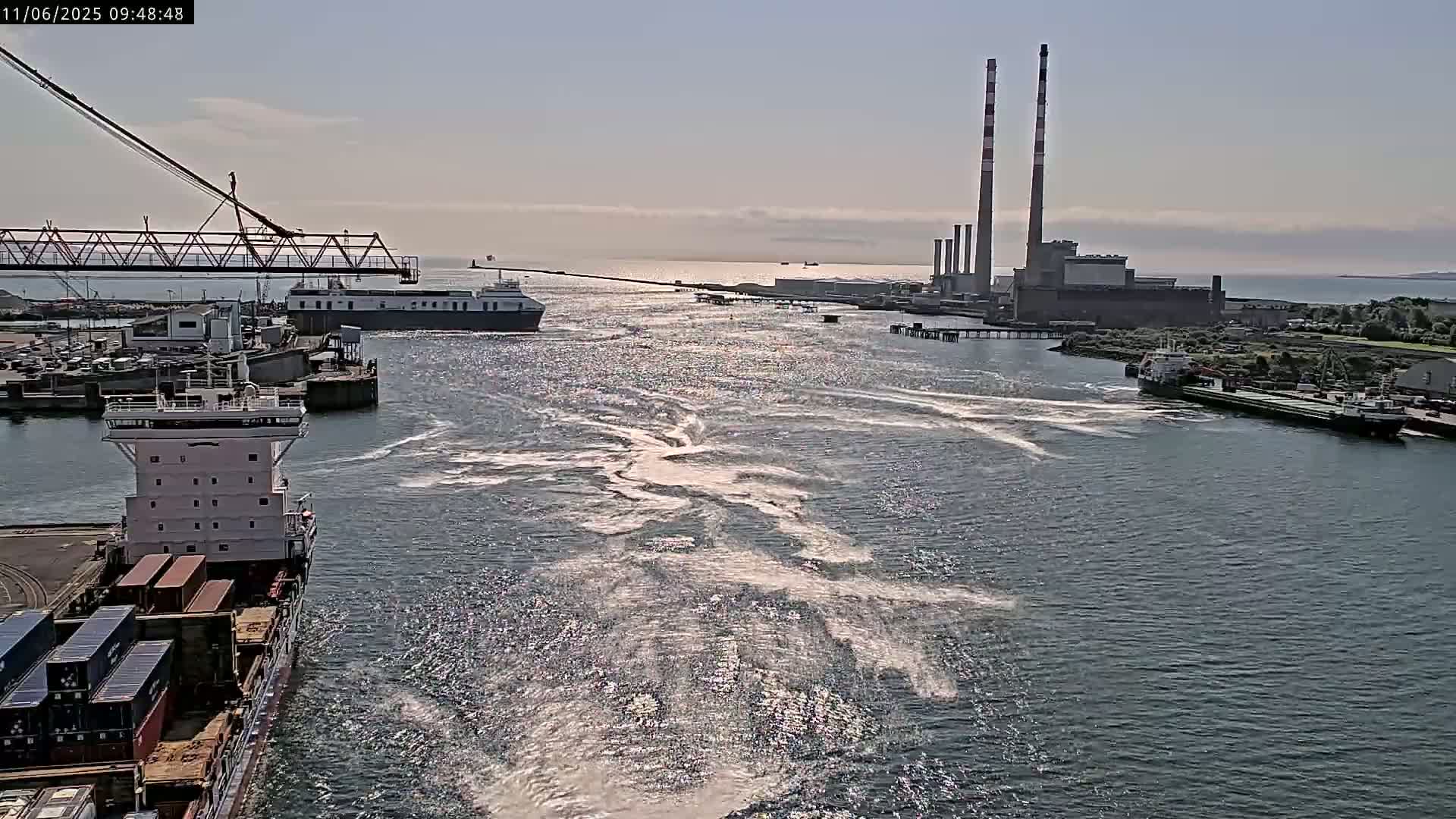 A container ship navigates a waterway past a ferry and toward a power plant under a mostly sunny sky.